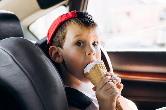 Close-up portrait of a child in a red baseball cap eating ice cream in a waffle cone while sitting in a car seat. Funny little boy riding in a car and looking at the camera