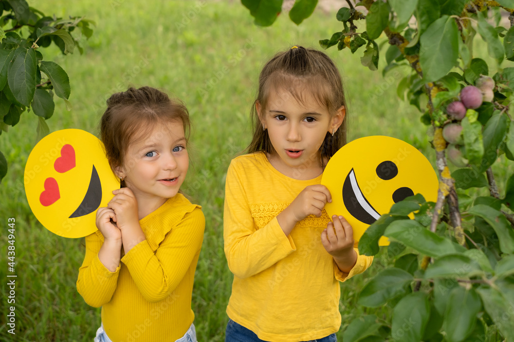 Wall mural The little sisters are holding the faces of happy emoticons in their hands. Two girlfriends in the park under an apple tree look with surprise and perplexity