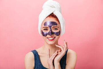 Front view of woman with towel and face mask. Smiling girl doing skincare routine isolated on pink background.