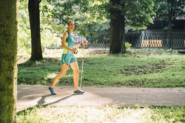 Obraz premium Nordic walking - full height middle-aged blond woman in sportswear working out in summer sunny city park.