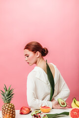 Pensive ginger woman with pineapple, bananas, garnets and kiwi. Studio shot of attractive girl with tropical fruits on pink background.