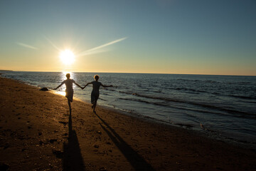 Fototapeta premium Two girls running hand in hand down the beach into the sea at sunset