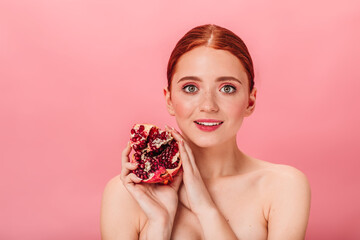Glad young woman holding garnet. Studio shot of naked girl with ripe pomegranate isolated on pink...
