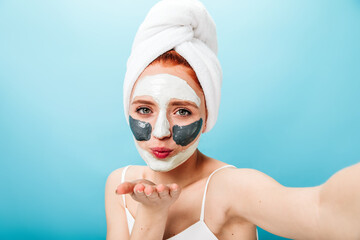 Glad caucasian girl with face mask sending air kiss. Beautiful lady doing skincare treatment and taking selfie.