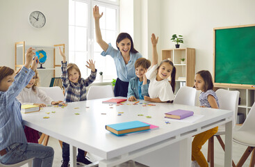 Happy clever enthusiastic school children and teacher raising hands. Confident preschoolers or first grade students sitting around big classroom table, learning letters, reading books and having fun