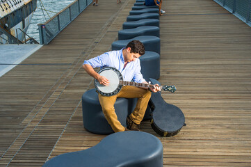 Dressing in a light blue shirt, dark yellow jeans and brown boot shoes, a young musician with an instrument box is sitting on a modern style bench, lowering his head and playing a banjo. - obrazy, fototapety, plakaty
