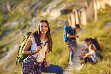 Smiling woman hiker with backpack looking at camera with group of friends hikers resting at...