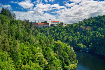 Fototapeta na wymiar Old Moravian castle, standing on a rock promontory above the river Dyje against a summer blue sky with white clouds, surrounded by green forest. Tourist area. Aerial view of Bítov castle, Czechia.