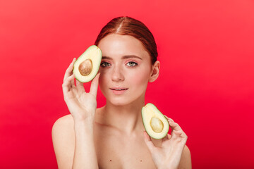 Good-humoured girl holding cut avocado. Studio shot of winsome ginger woman isolated on red...
