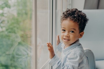 Cute mixed race little curly haired boy in light blue colored shirt next to large window