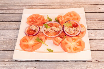 Salad of sliced peeled tomatoes, cherry tomatoes, red onion rings and canned tuna on white square plate