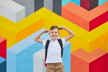 Cheerful schoolboy showing muscles against colorful wall