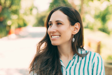 Portrait of young woman with brown hair in fashionable clothes smiling and looking away outdoor