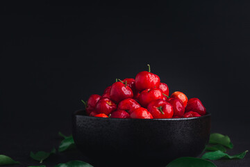 Red acerola cherries fruit in a ceramic bowl with a black background. High vitamin C and antioxidant fruits. Close-up. Side view. Space for text. Concept of healthy fruits