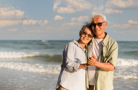 Asian senior couple tourist is hand in hands, embrace and dancing on tropical sea beach in summer holiday.
