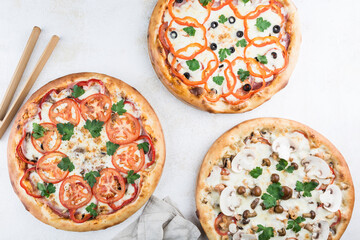 Three pizzas with different fillings of sausage, ham, mushrooms, bell pepper, tomatoes, Mozzarella cheese and parmesan on a light background.