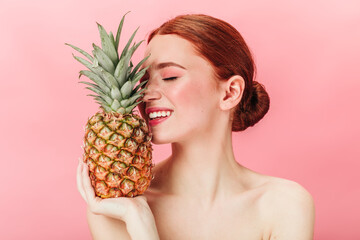 Stunning ginger girl holding pineapple with closed eyes. Studio shot of gorgeous caucasian woman with fruit posing on pink background.