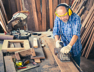 Happy senior carpenter using an electric planer with a wooden plank in the carpentry workshop. Manufacture of wood products..