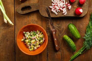 Okroshka in a clay plate on a table on a wooden table next to vegetables on boards and a knife.