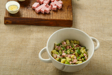 Okroshka in a clay plate on the table on a tablecloth next to an egg sausage and cucumbers on boards.