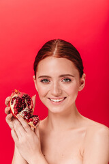 Dreamy ginger woman holding garnet and laughing. Happy nude girl posing with pomegranate on red background.