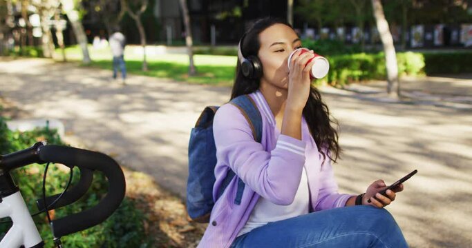 Asian woman wearing headphones listening to music and drinking takeaway coffee