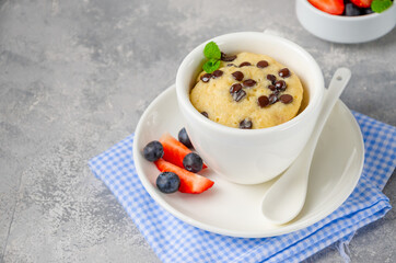 Mug cake with chocolate chips, fresh berries and cream on a gray concrete background. Copy space.