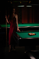 Young curly girl posed near billiard table. Sexy model at red dress.