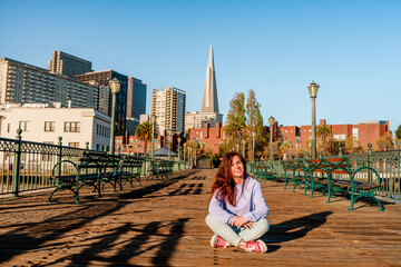 A young woman in jeans sits on a wooden floor on a pier in the morning with a view of downtown and the Transamerica Tower in San Francisco