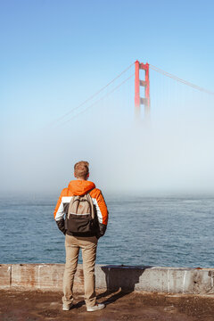 A young man with a jacket stands on the embankment and looks at the fog covering the Golden Gate Bridge in San Francisco, an incredible landscape