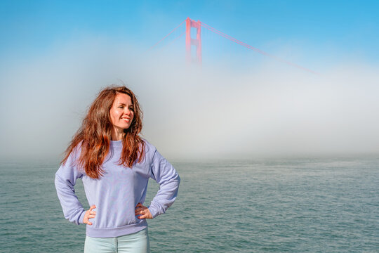 A young woman stands on the embankment overlooking the Golden Gate Bridge covered with fog in San Francisco, an incredible landscape