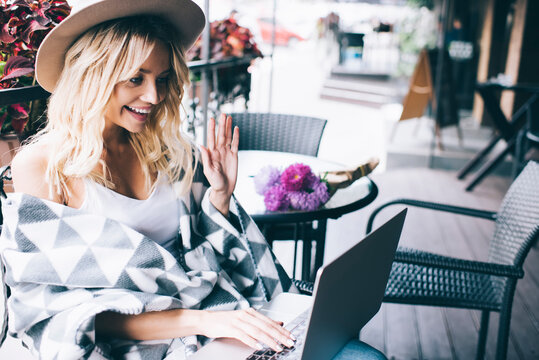 Happy Caucasian woman in trendy hat browsing website for making contact calling via laptop application, cheerful hipster girl waving while talking during web conversation on digital netbook