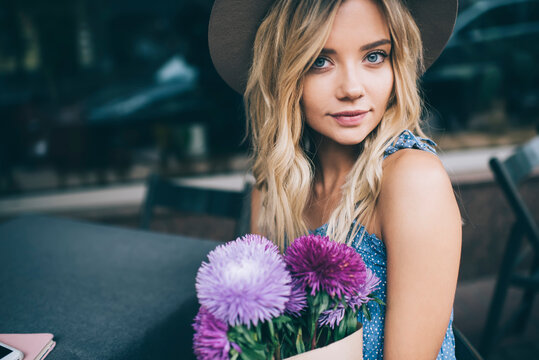 Portrait of attractive Caucaisan woman with beautiful violet flowers looking at camera during weekend pastime in street cafe, young hipster girl holding present of floristic delivery and posing