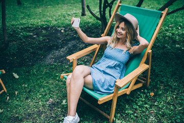 Carefree female blogger enjoying time for creating selfie content using front camera on cellular device, happy hipster girl resting in park making influence video vlog via smartphone application