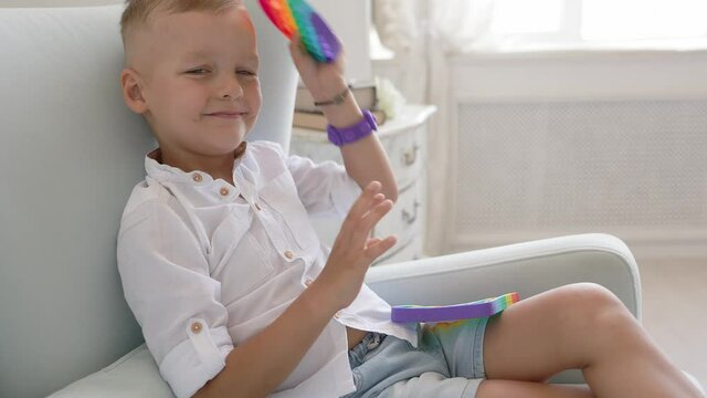 Portrait blonde boy with pop it sensory toy different forms. Boy presses colorful rainbow squishy soft silicone bubbles in children's playroom. Stress and anxiety relief. Trendy fidgeting game.