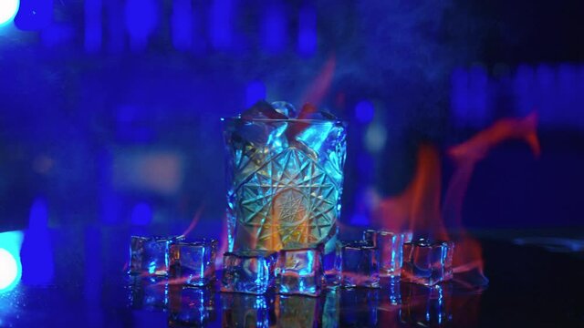 Bartender sets fire to alcohol cocktail, blue background. Flames in cocktail glass in slow motion, burning ice in alcohol drink, barman makes drink.