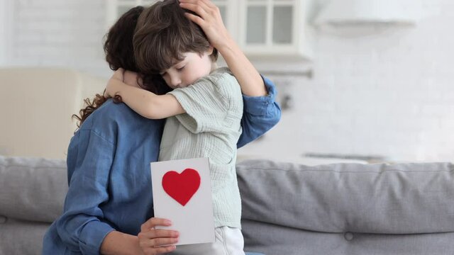 Cute small caucasian boy congratulates mom with holiday, tender embrace young female parent holding handmade postcard gift to happy mum birthday or mother day. Preschool boy and loving mummy hugging