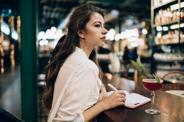 Contemplative female student with education textbook and alcohol beverage thinking about information in restaurant, Caucasian woman with notepad planning organisation during cocktail time in bar