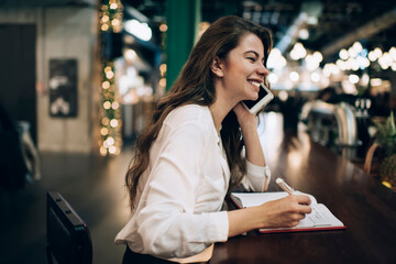 Cheerful female student with notepad for learning and studying making positive cellphone communication and laughing, joyful hipster girl with education copybook rejoicing while phoning via app