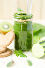 Healthy green smoothie with spinach, kiwi and orange in a retro jar on light wooden table. Integral biscuits and cucumber in the foreground, kiwi in the background.