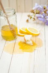 Honey dipper in honey with honeycomb, lemon, flowers and jar on light wooden table.