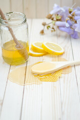 Honey dipper in honey with honeycomb, lemon, flowers and jar on light wooden table.