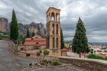 Naklejka premium A church with a tall elegant bell tower on a hill in front of huge rocky mountains above a town, Kalambaka, Meteora, Greece