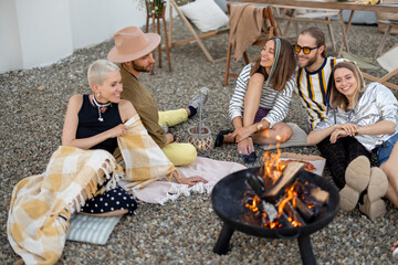 Young stylish friends sitting together by the fireplace outdoors. Talk and have fun during the...