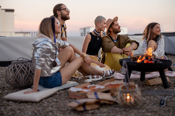 Group of young stylish friends sitting together by the fireplace, having a picnic on the rooftop...