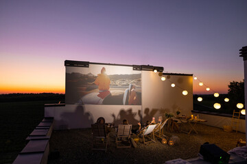 Small group of people watching movie on the rooftop terrace at sunset. Open air cinema concept. Romantic leisure and entertainment on the roof of a country house