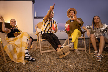 Obraz premium Young people watching football match or some film with popcorn outdoors on a rooftop terrace at night. Open air cinema concept