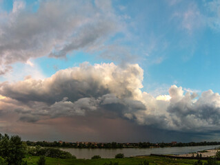 powerful thunderstorm front with red-dark clouds and blue sky over a flat river - panorama on a summer day