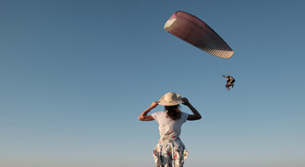 Slim girl stands and looks at the paraglider. Woman stands on a hill holding a hat in her hands and...