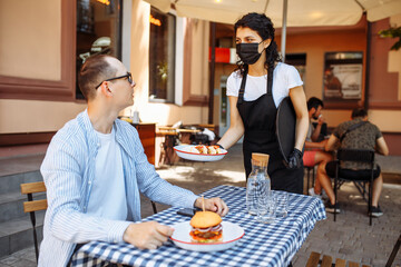 Happy waitress serving food and drinks to her customer while wearing protective face mask and gloves due to coronavirus epidemic.